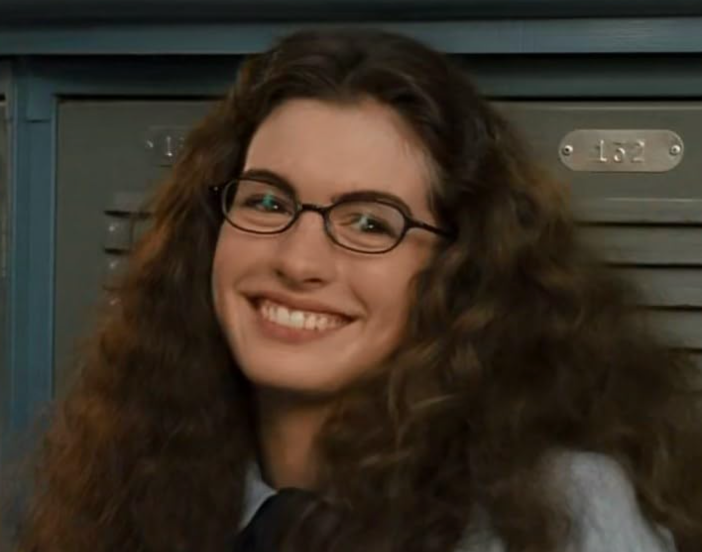 A smiling person with curly hair and glasses stands in front of lockers, wearing a shirt and tie