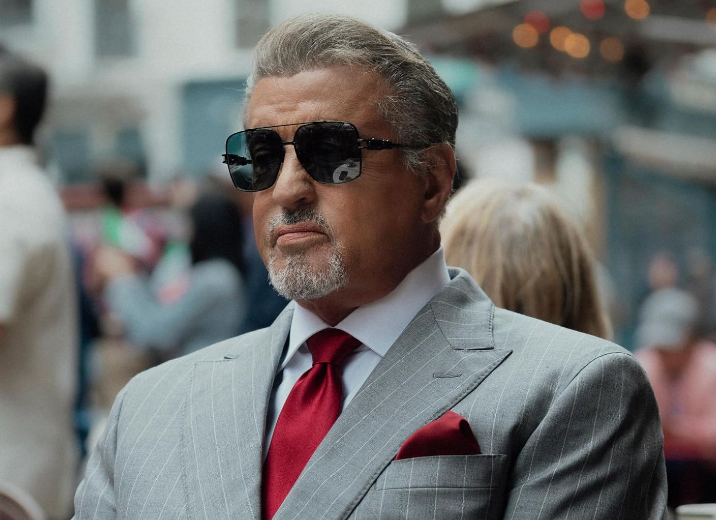 Person in a sharp suit, wearing sunglasses and sitting outdoors with a serious expression