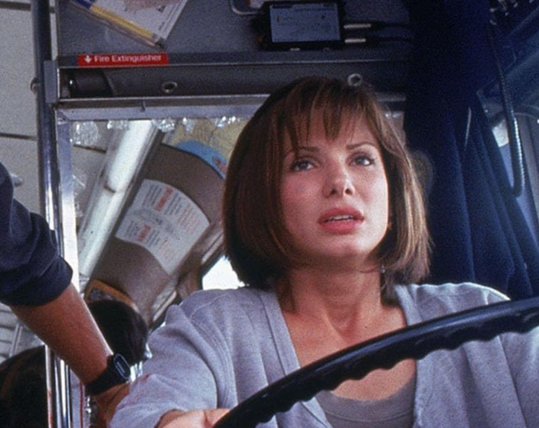 Person gripping steering wheel inside a bus, looking concerned. Nearby, another person's hand is visible wearing a watch. Bus interior details seen
