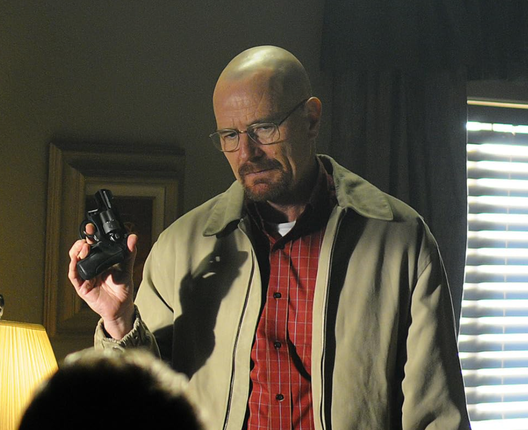 A bald man in glasses and a jacket holds a gun, looking intently at something off-camera in a dimly lit room