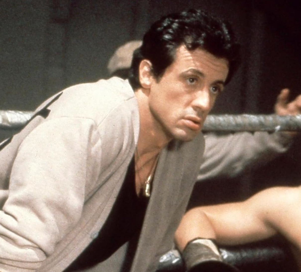 A man in a boxing ring leans on the ropes, appearing contemplative and focused
