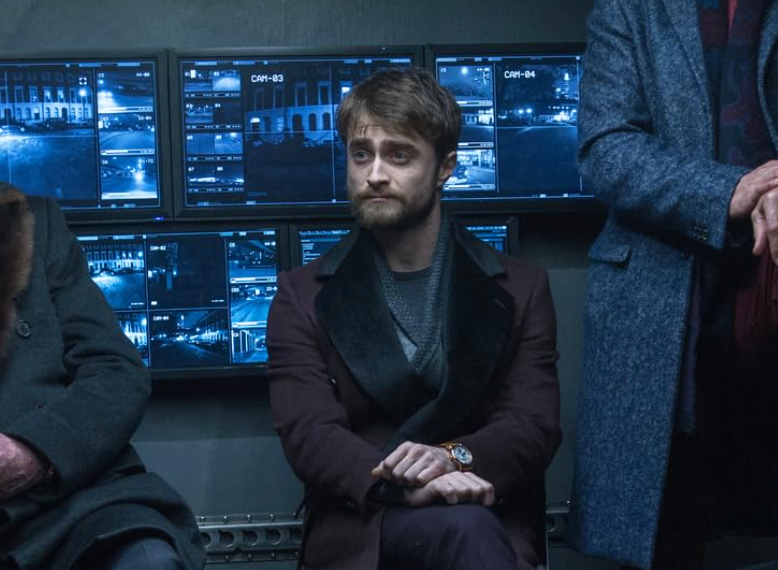 A man sits with crossed legs in front of several security monitors, wearing a dark coat and a watch