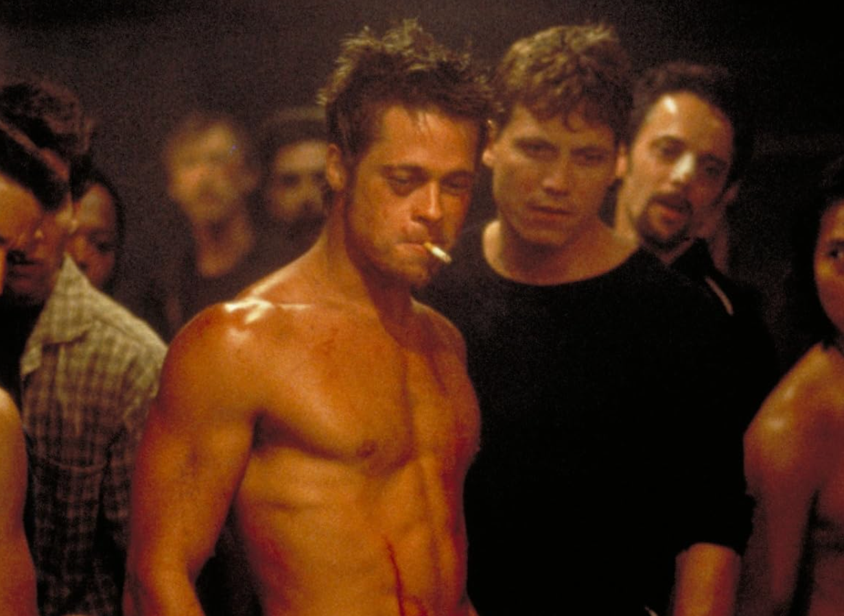 Shirtless man with a cigarette stands confidently in a dim, crowded room. Others, also with intense expressions, surround him closely
