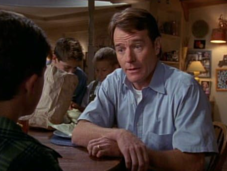A man in a casual shirt talks seriously to a young boy at a dining table while other children are in the background