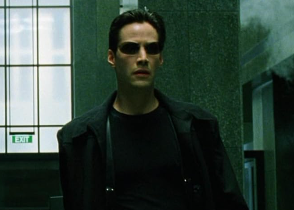 Person wearing a black coat and sunglasses in a futuristic setting, with a determined expression walking indoors