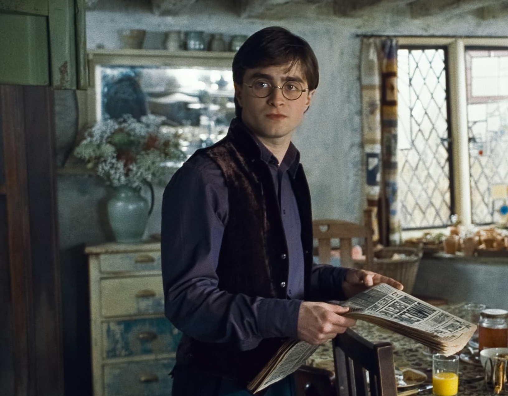 Person with glasses in a rustic kitchen, holding a newspaper and looking curious. Background includes floral arrangements and kitchenware