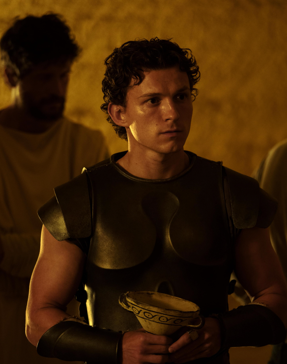 Person in ancient armor holds a decorated bowl, standing in a dimly lit area with others in the background