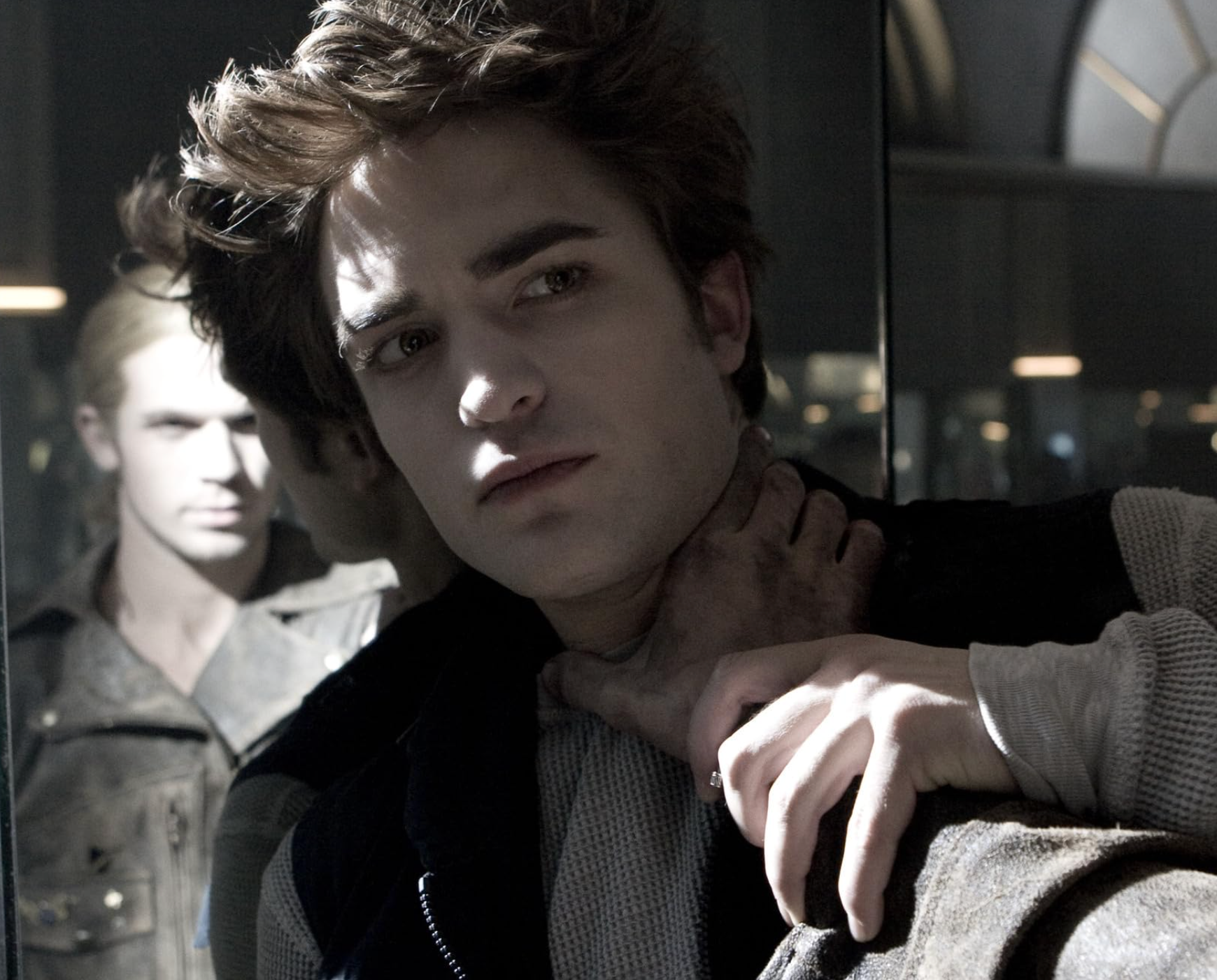 A pensive man with styled hair is held by another in a tense scene, reflected in a mirror. The setting is dimly lit, suggesting drama or conflict