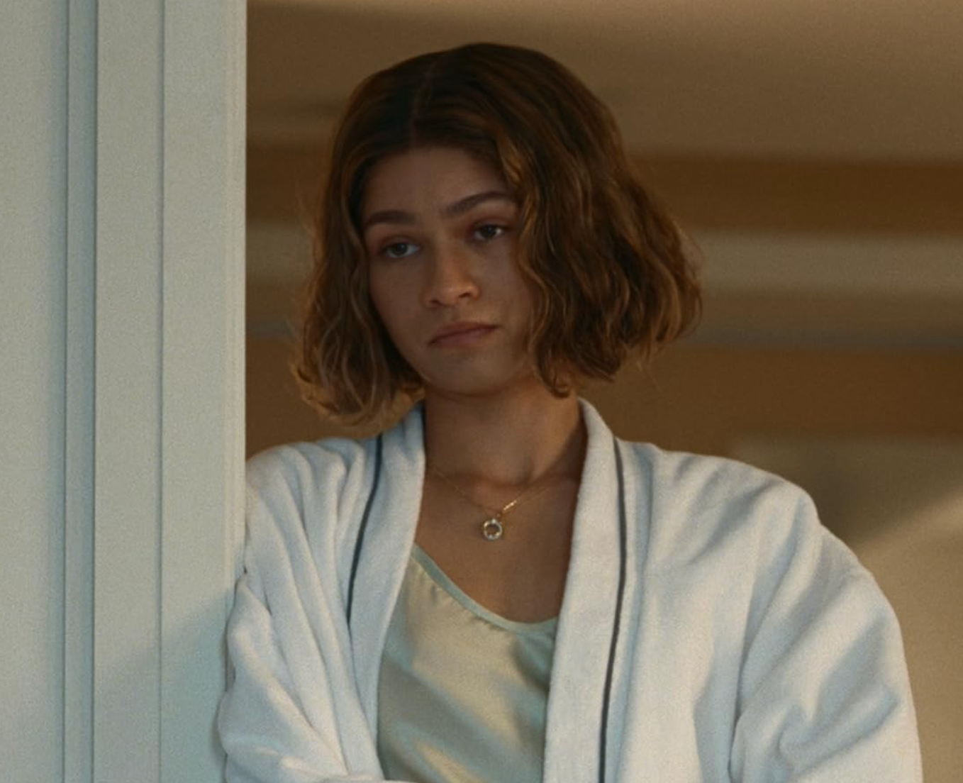 Person with short wavy hair in a white robe looks contemplative while standing indoors