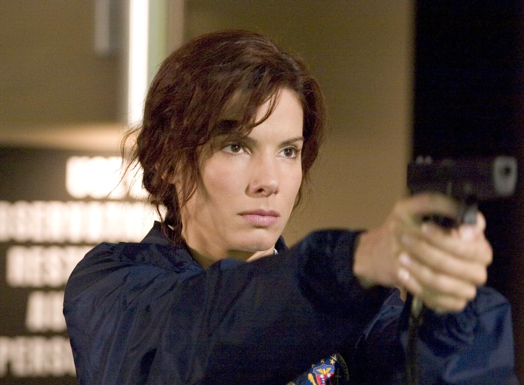 Person in a focused pose holding a gun, wearing a dark jacket, appears to be from a scene in a film or TV show