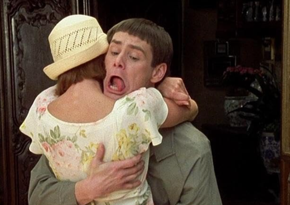 A man with a comedic expression embraces a woman in a floral dress and hat