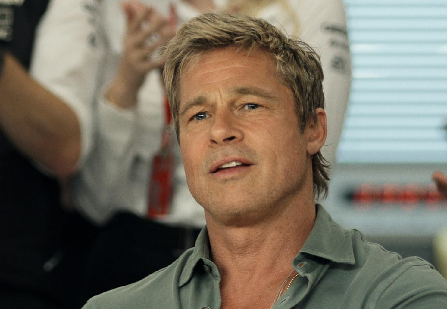 A person with short hair in a casual shirt is seated, with others clapping in the background
