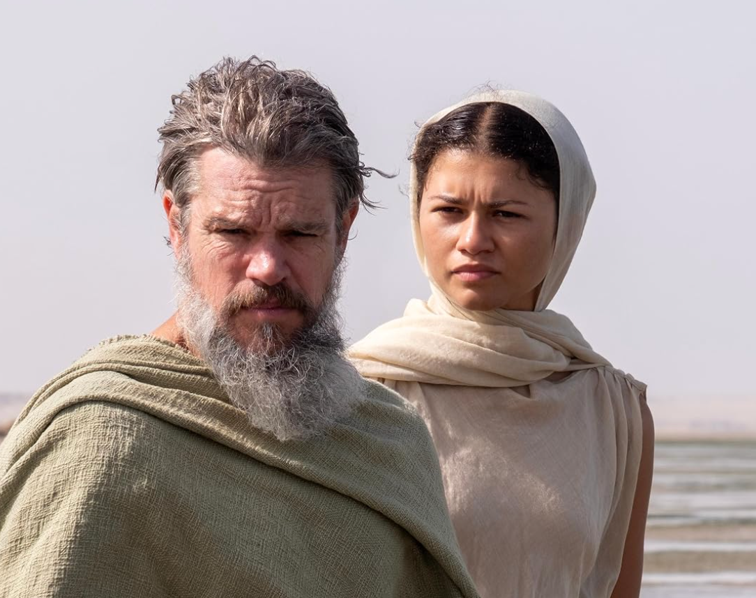 Two people in desert attire stand outdoors. One has a beard and wears a cloak; the other wears a headscarf and looks intently ahead