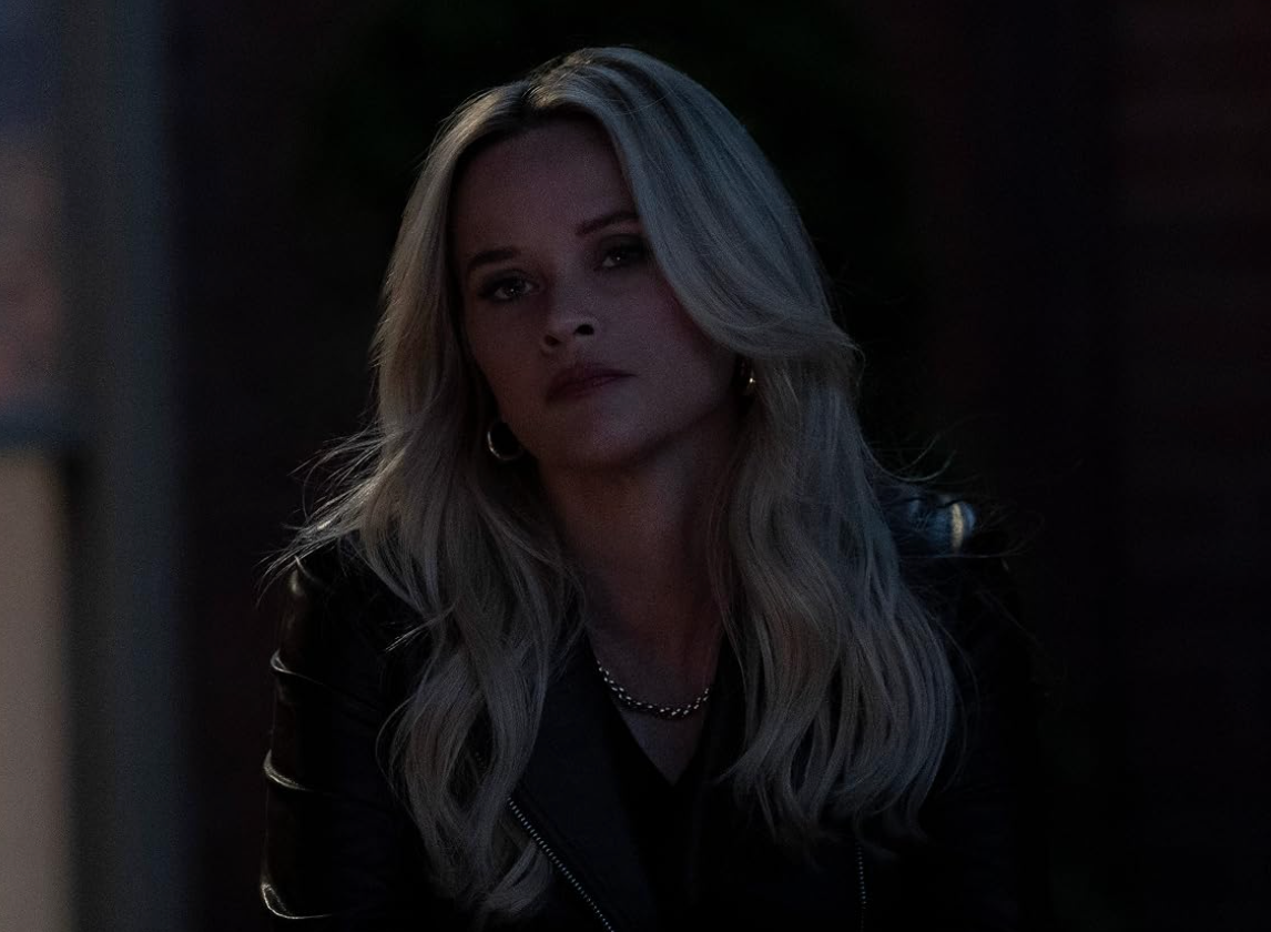 A person with long hair sits in dim lighting, wearing a leather jacket and a necklace, gazing thoughtfully