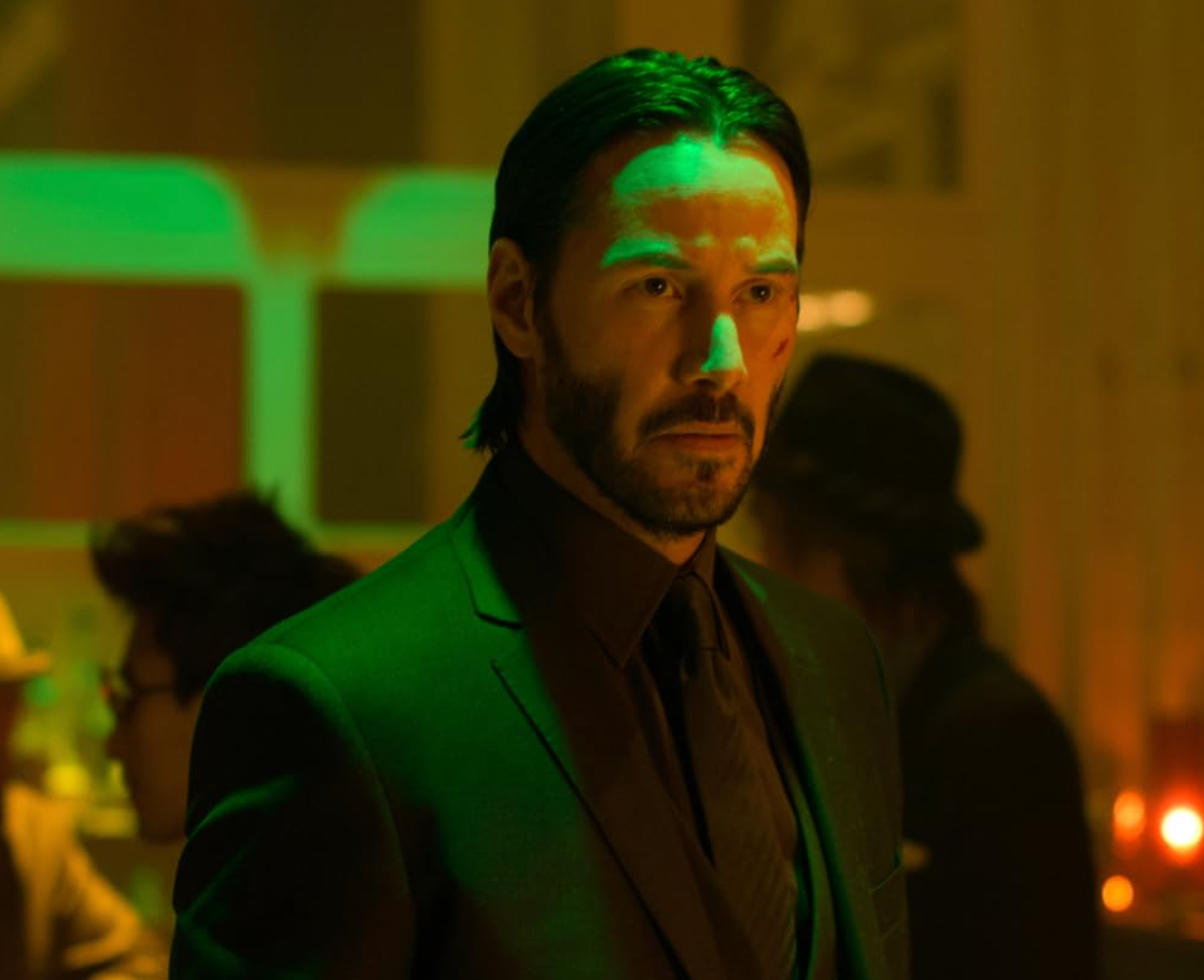 A person in a dimly lit setting wearing a tailored suit, with visible cuts on their face, looks pensively ahead