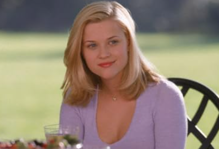 A woman with shoulder-length blonde hair sits at an outdoor table, wearing a V-neck top and smiling gently