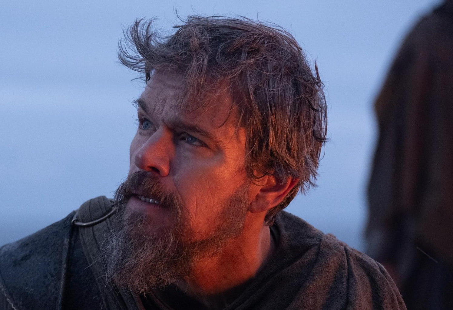 A bearded man with tousled hair looks thoughtfully into the distance, wearing a rugged outdoor outfit