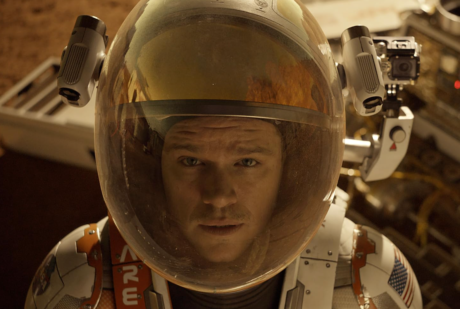 A person in a space suit with a clear helmet visor looks intently at the camera, set against a sci-fi backdrop