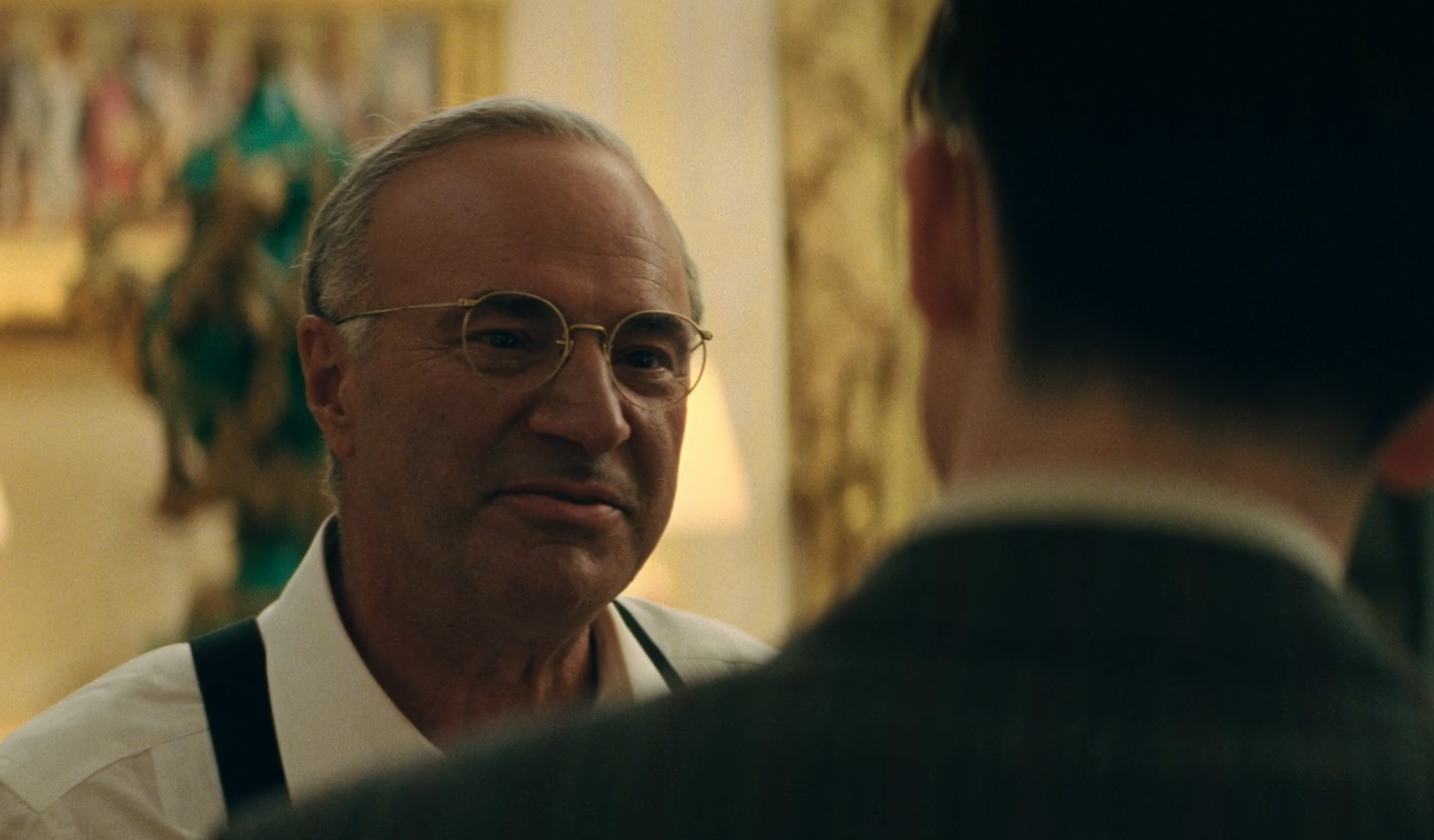 Two people engaged in conversation in a room with ornate decor. One person is wearing glasses and suspenders