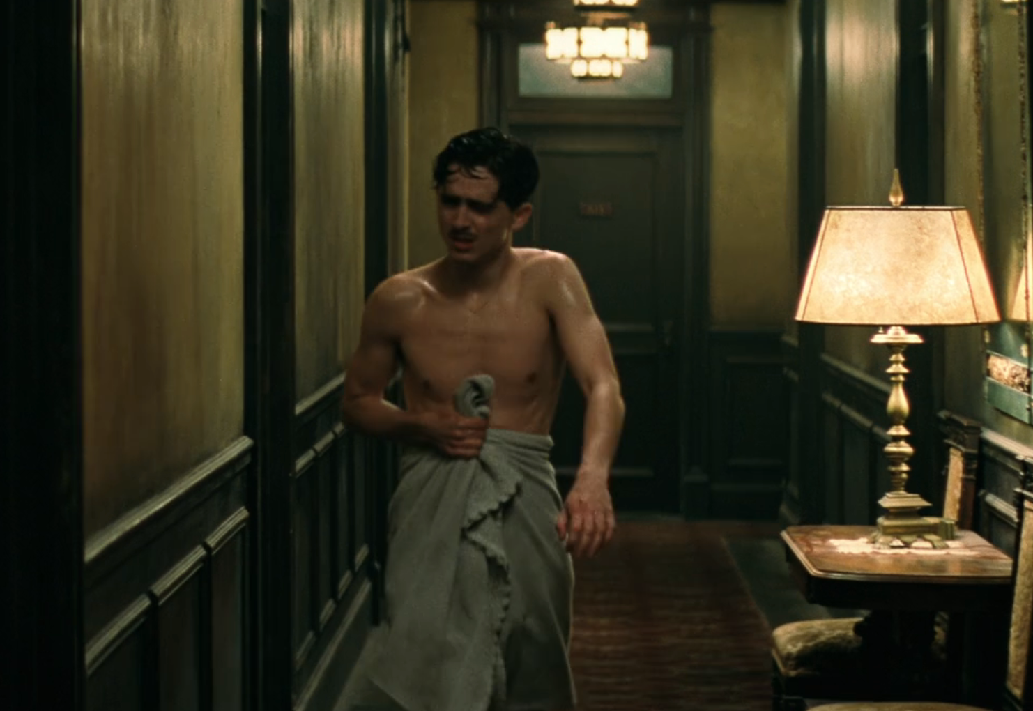 A man walks down a dimly lit hallway wrapped in a towel, appearing wet and thoughtful