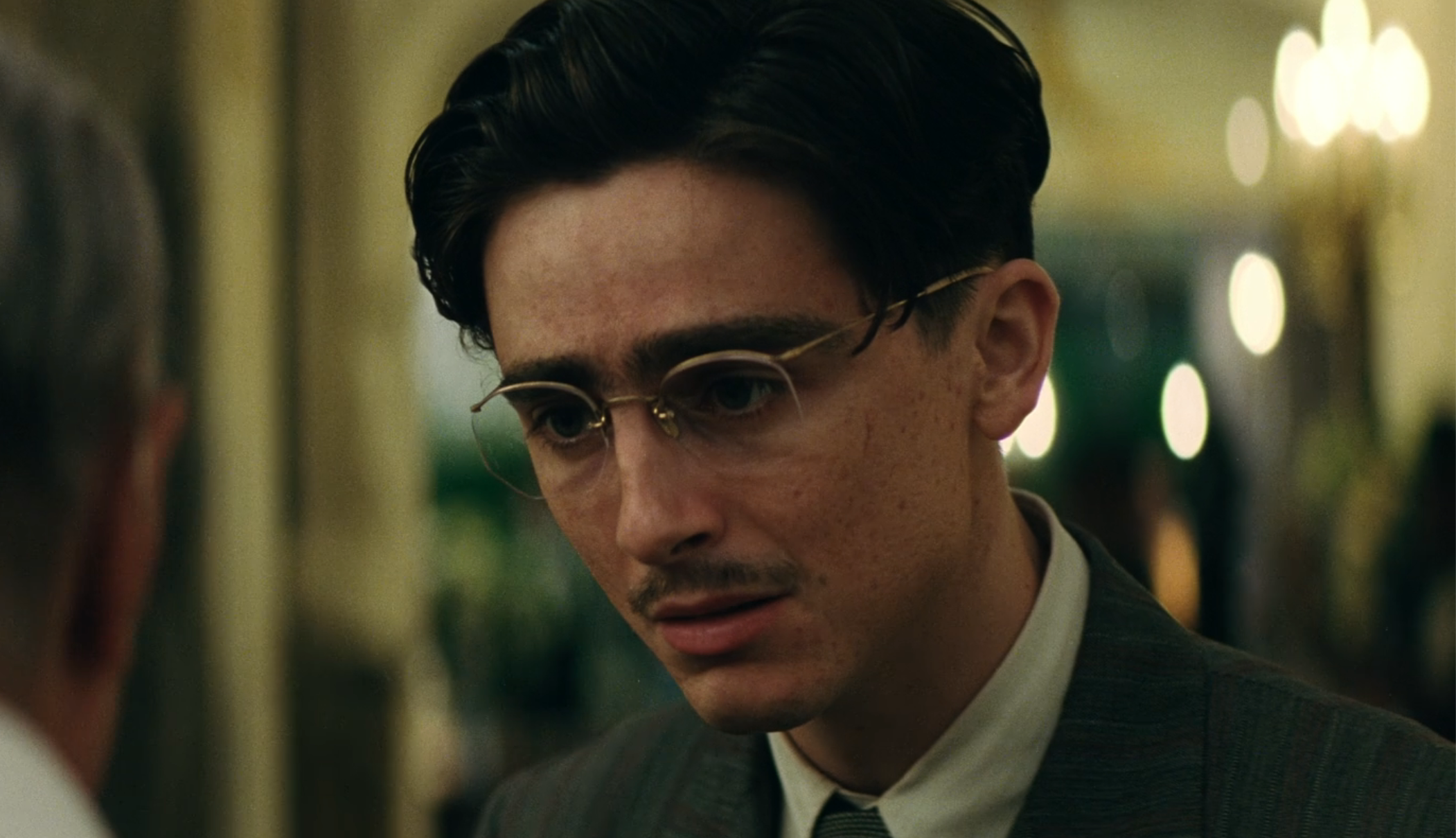 A person in a suit and glasses looks intently at someone off-camera in a formal setting
