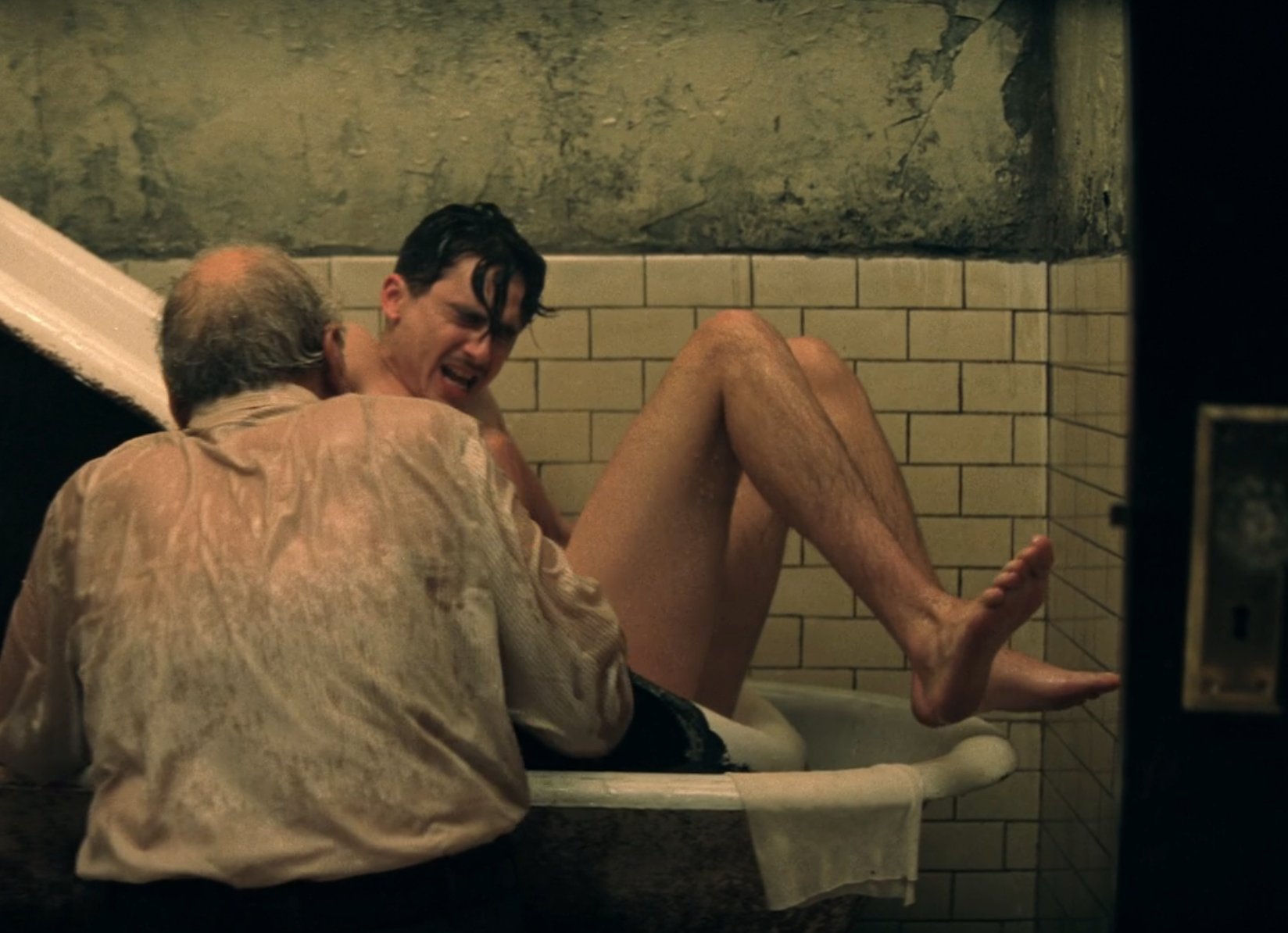 Two men in an old bathtub, one sitting inside, the other assisting. Worn tiles and peeling walls suggest an aged or gritty setting