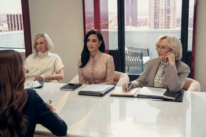 Three people sit at a table in business attire, engaged in a meeting. One wears a white blouse, one a light dress, and one a tan suit