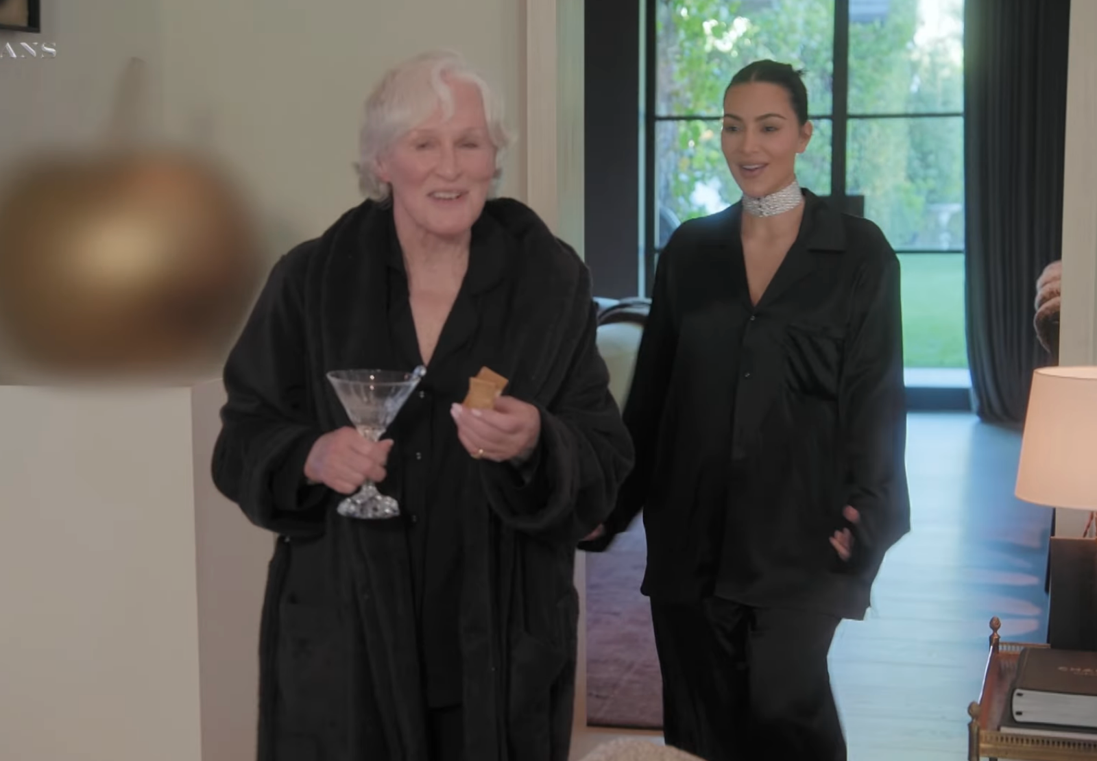 Two individuals in a home setting; one holds a martini and cracker, wearing a robe, while the other in sleek attire and choker looks on smiling