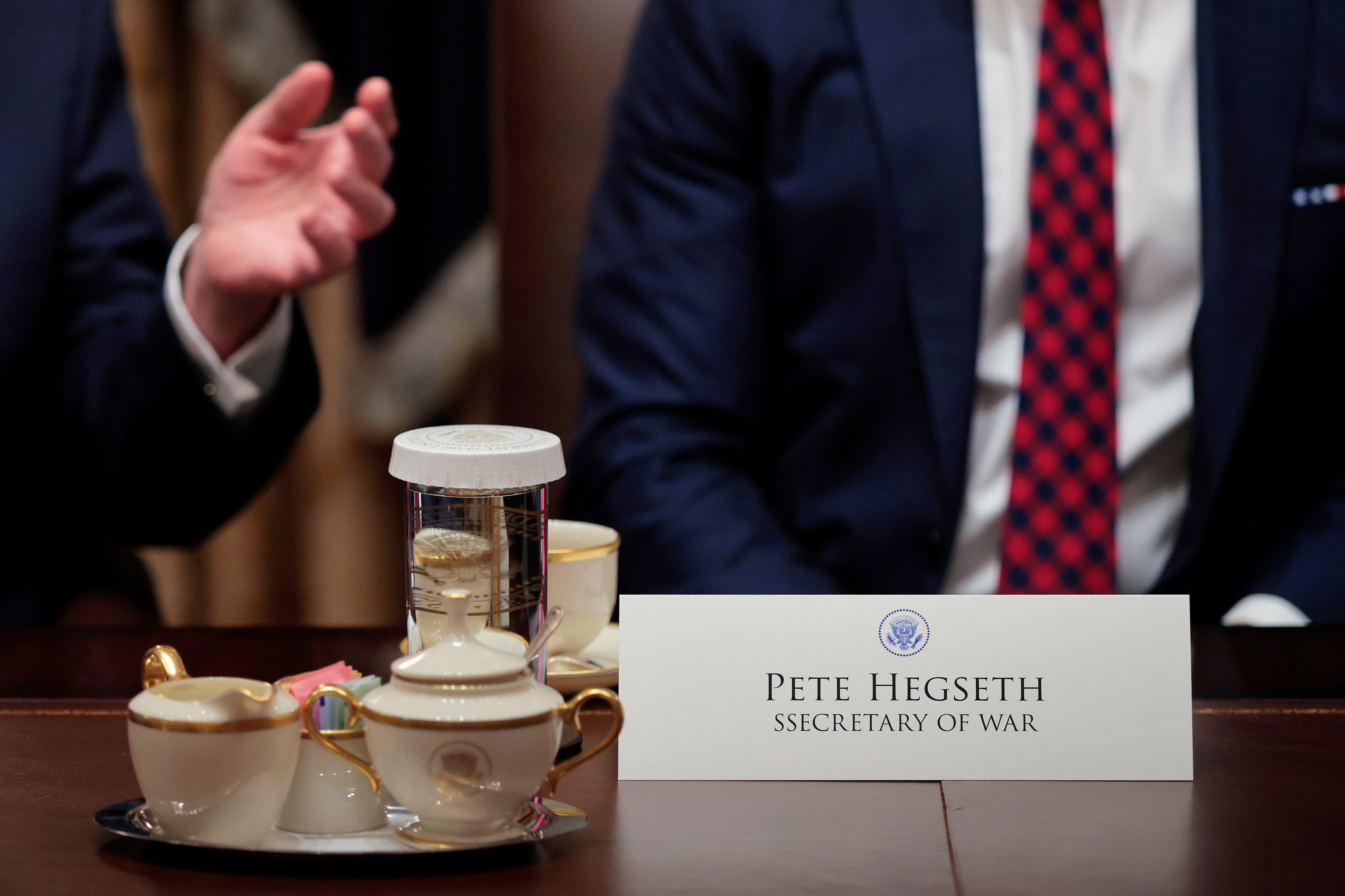 To personer i dress sitter ved et bord med bordkort som leser «Pete Hegseth, krigsminister,»; og et tesett med en tallerken med søtsaker foran