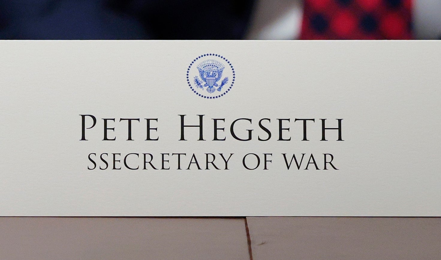To personer i dress sitter ved et bord med bordkort som leser «Pete Hegseth, krigsminister,»; og et tesett med en tallerken med søtsaker foran