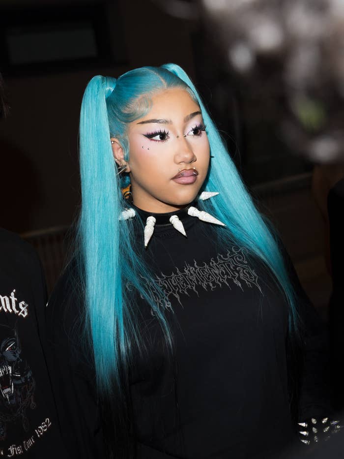 Person with long blue pigtails and dramatic makeup wears a black hoodie with graphic text and a spiked choker