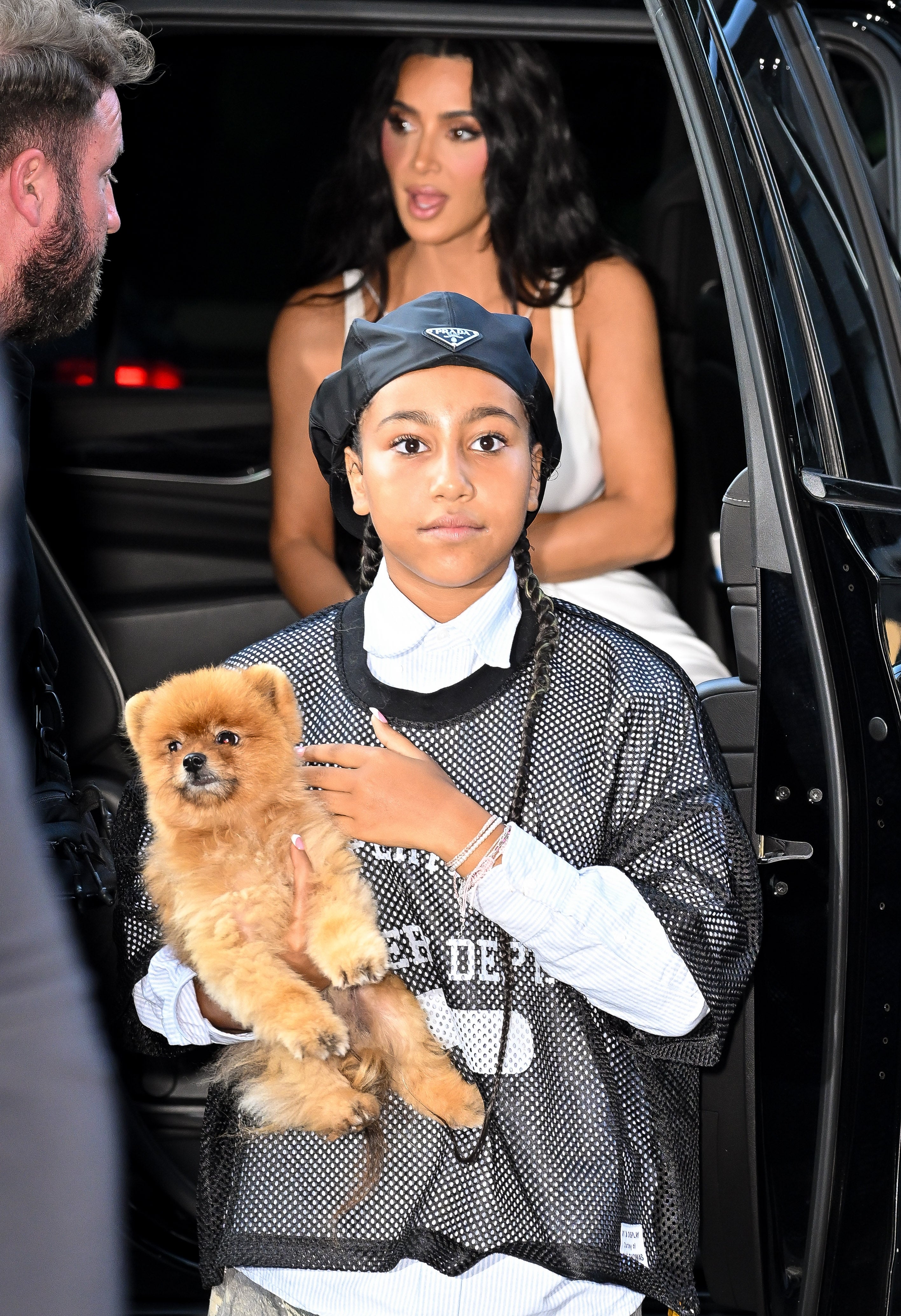 A person in an elegant gown exits a car while another holds a small fluffy dog, dressed in a mesh sports jersey and a bandana