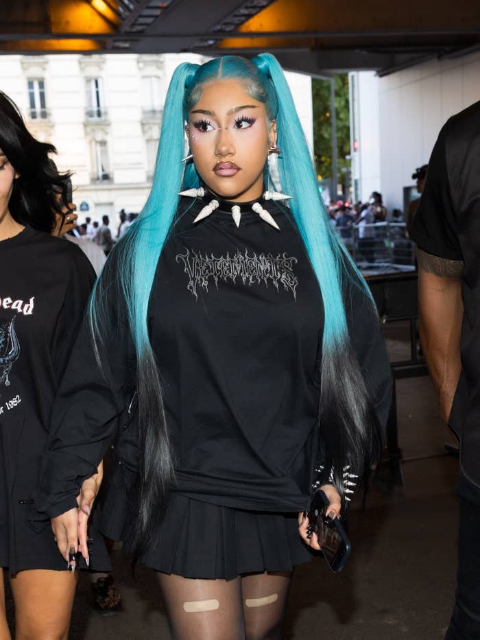 Person in edgy fashion with long hair, graphic top, spikes necklace, and pleated skirt at an outdoor event