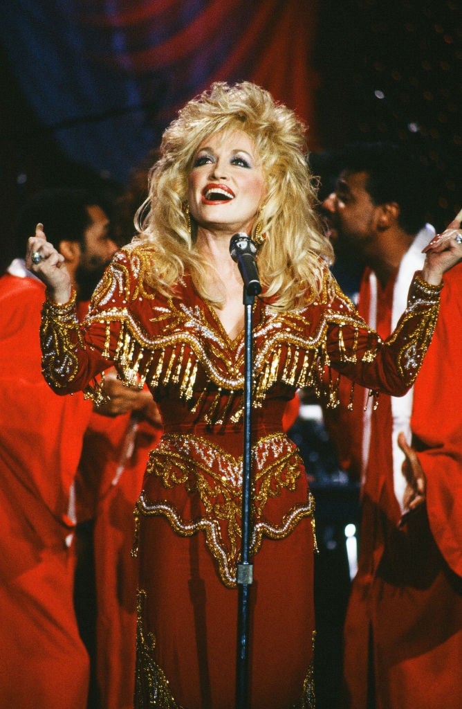 Singer performing on stage, wearing a glittering, fringed gown. Backup singers in matching outfits are in the background