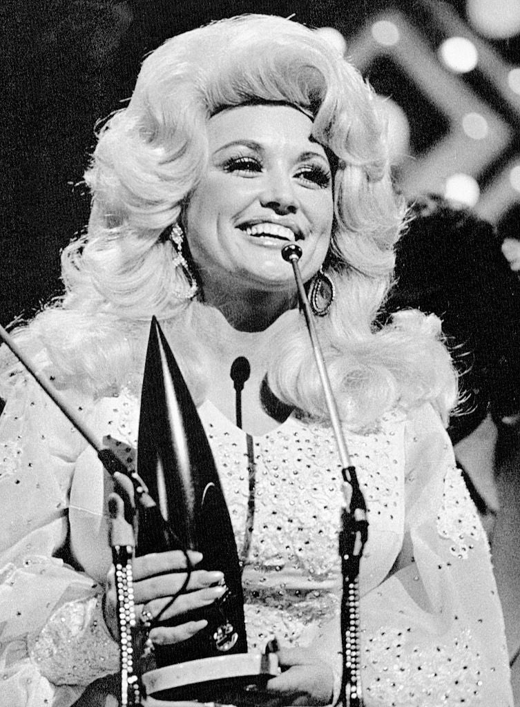 Person on stage holding a music award, wearing a sparkling, sequined dress with voluminous hair, smiling at the audience