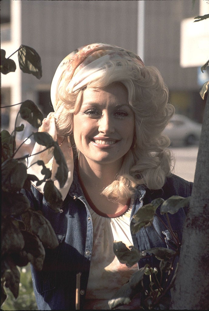 Person with long, wavy hair smiles while peeking through leaves. Wearing a denim top over a light blouse