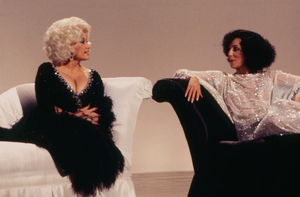 Two people in glamorous outfits sit on elegant couches, engaged in conversation. One wears a sparkling gown, the other a black dress with feather accents