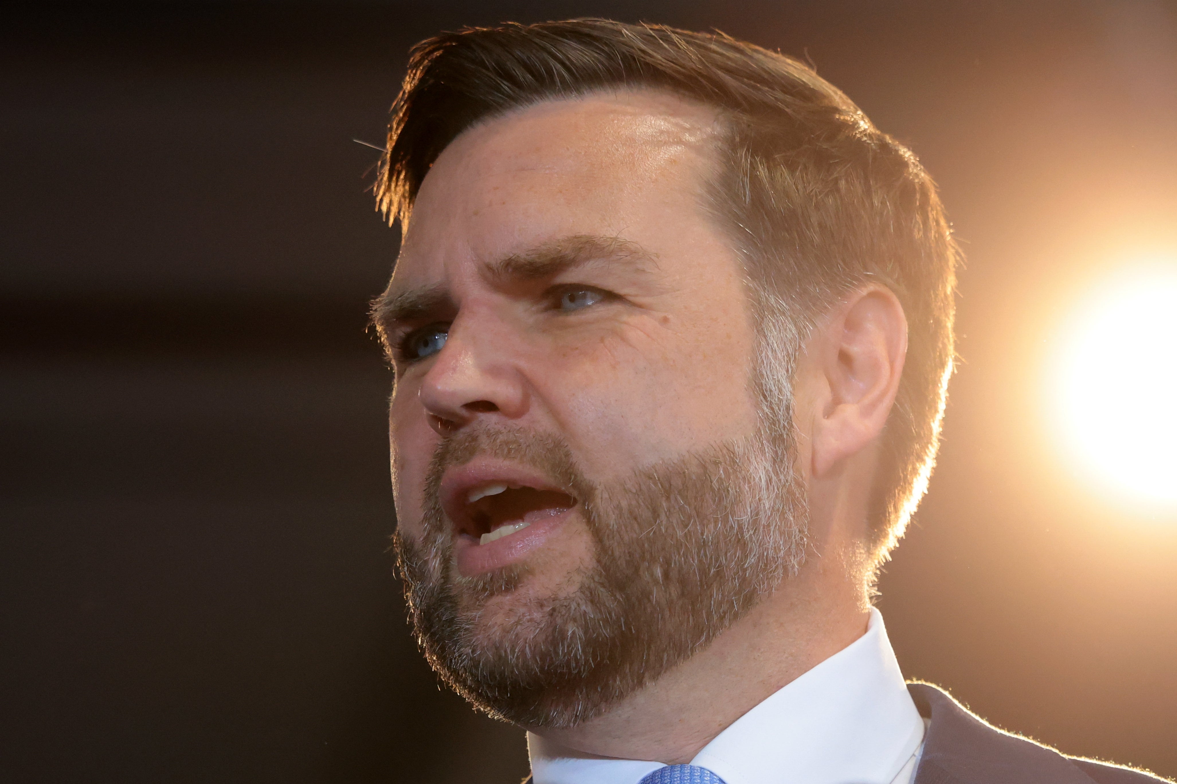 En mann med kort hår og skjegg snakker lidenskapelig på et arrangement, opplyst av et sterkt lys bakfra