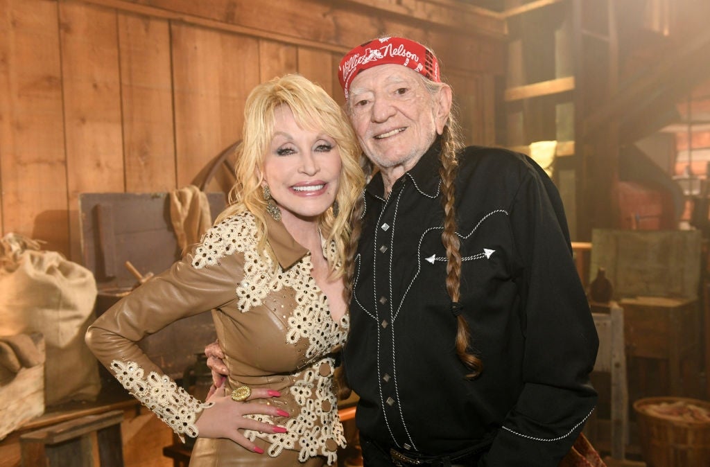 Two people smiling in rustic setting; one in a lace-trimmed leather outfit, the other in a Western shirt and bandana
