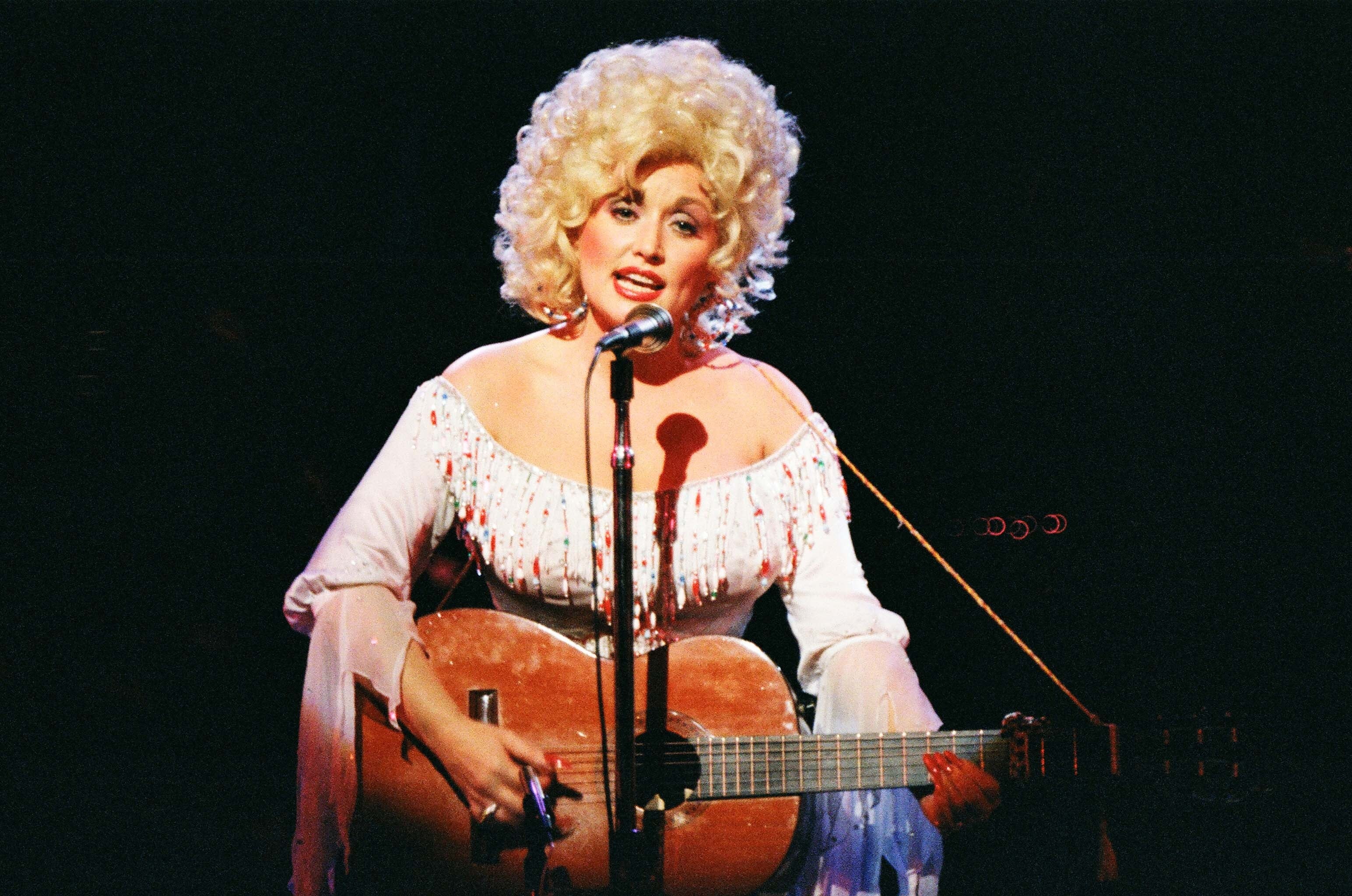 Person with voluminous curly hair performs on stage, wearing a sequined outfit and playing an acoustic guitar