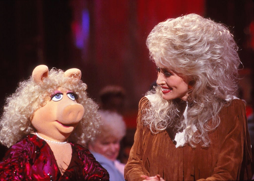 A puppet and a person smile at each other on a stage. The puppet has a pearl necklace; the person wears a fringed outfit