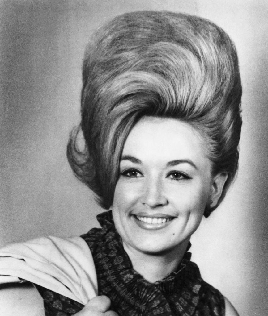Person smiling with a large, high-volume hairstyle, wearing a patterned sleeveless top and draped fabric over shoulder