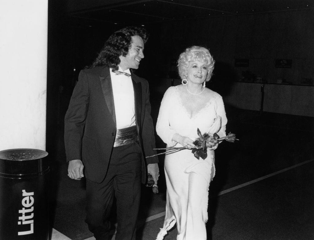 A man in a tuxedo walks with a woman wearing a glamorous long dress and holding flowers at an event