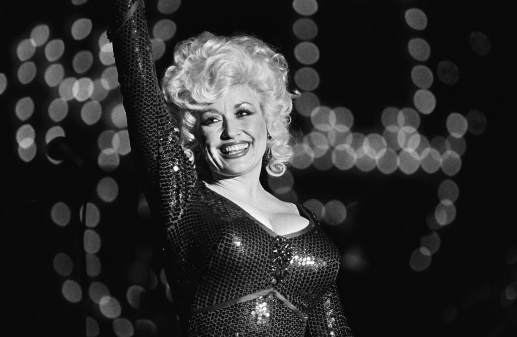 A person with voluminous hair smiles on stage, wearing a glittering sequin dress, against a backdrop of blurred lights