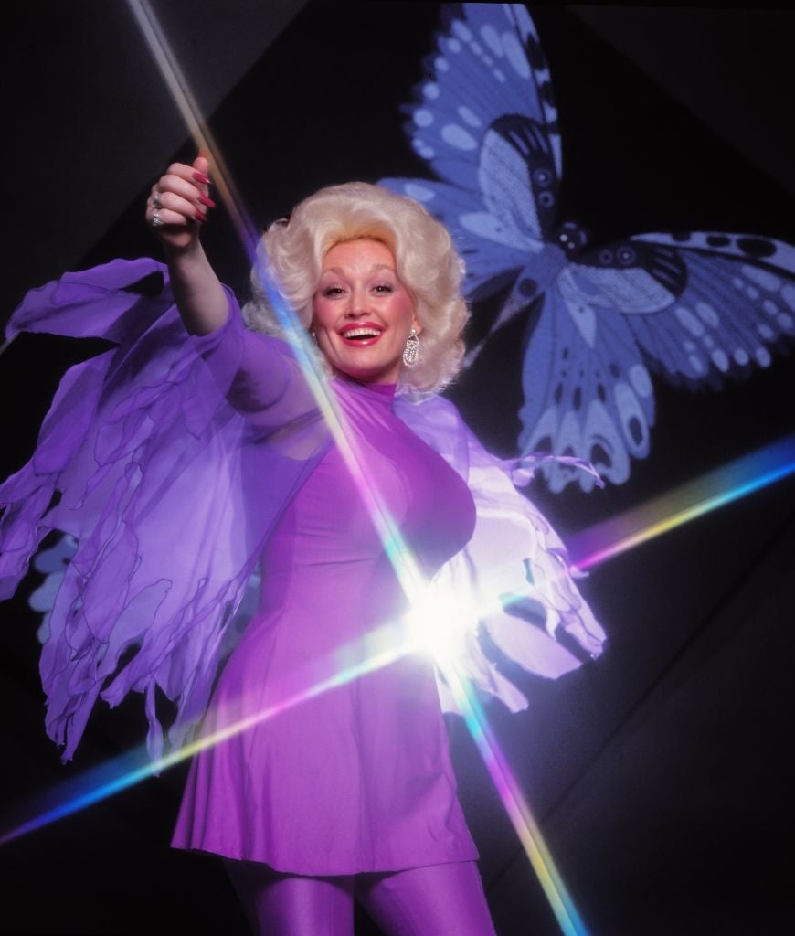 Person in butterfly-themed outfit with large wings poses joyfully, holding a wand. Bright light and butterfly backdrop emphasize a whimsical feel
