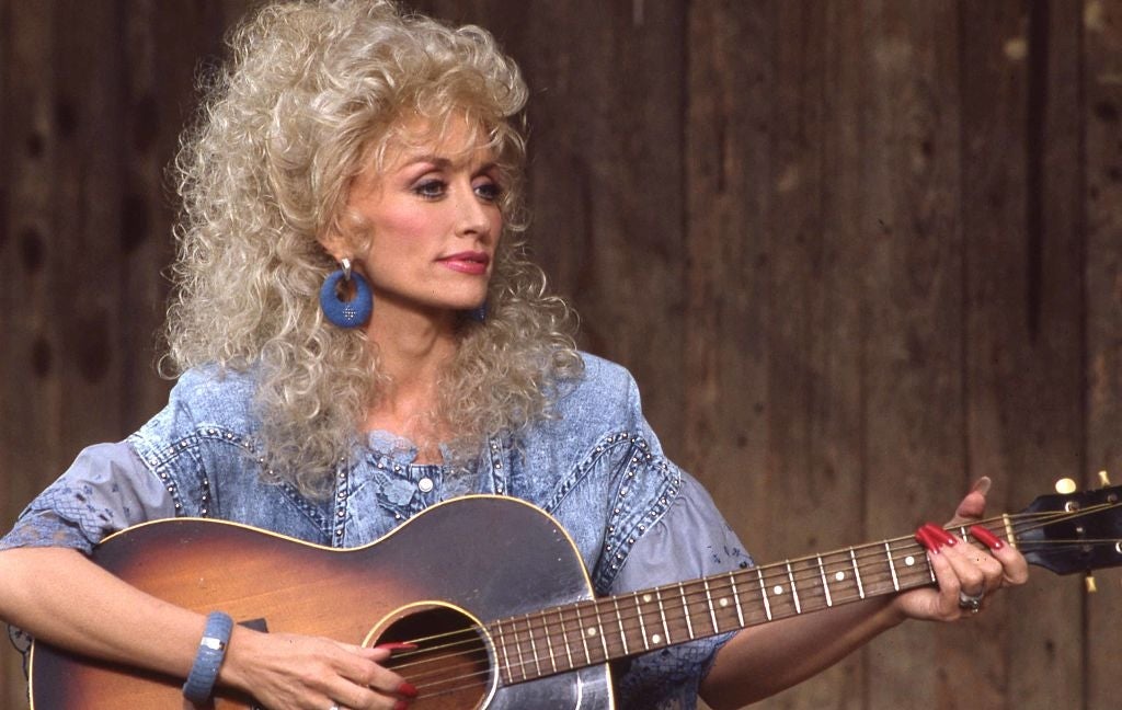Person with curly hair playing an acoustic guitar, wearing a denim outfit and statement earrings, focused intently on playing music