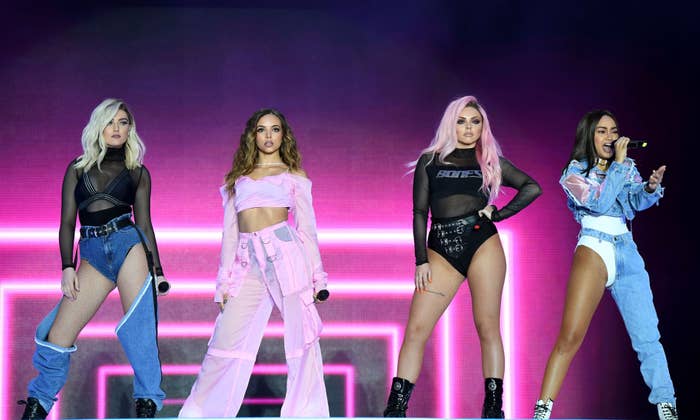 A music group performs on stage, each wearing unique, bold outfits including crop tops, high-waisted pants, and shorts, with dynamic poses and a neon backdrop