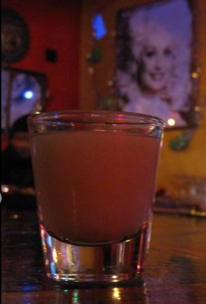 A glass filled with a yellowish drink sits on a bar with a blurred portrait of a woman in the background