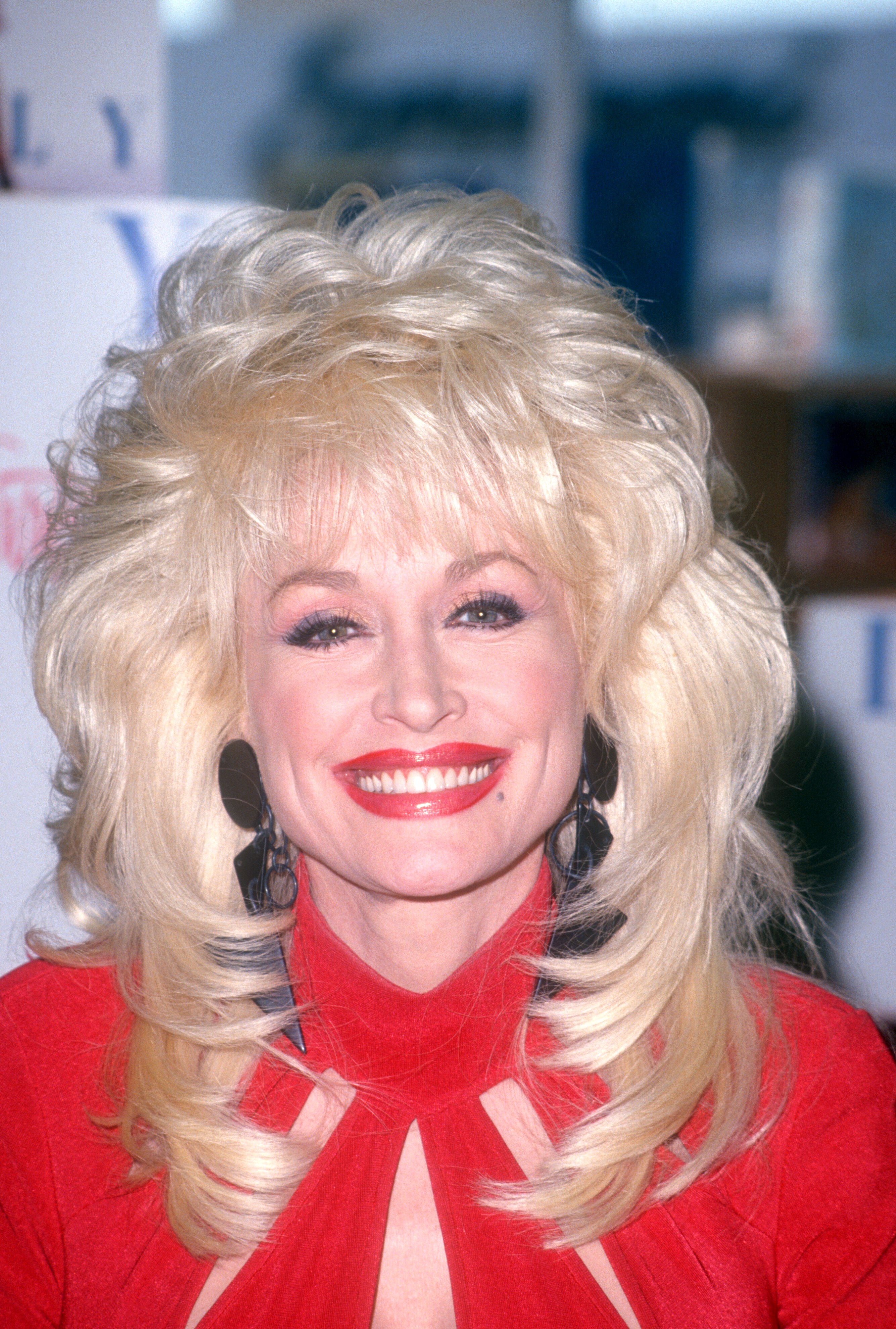 Person with voluminous blond hair, wearing a high-neck red dress, smiling at an event