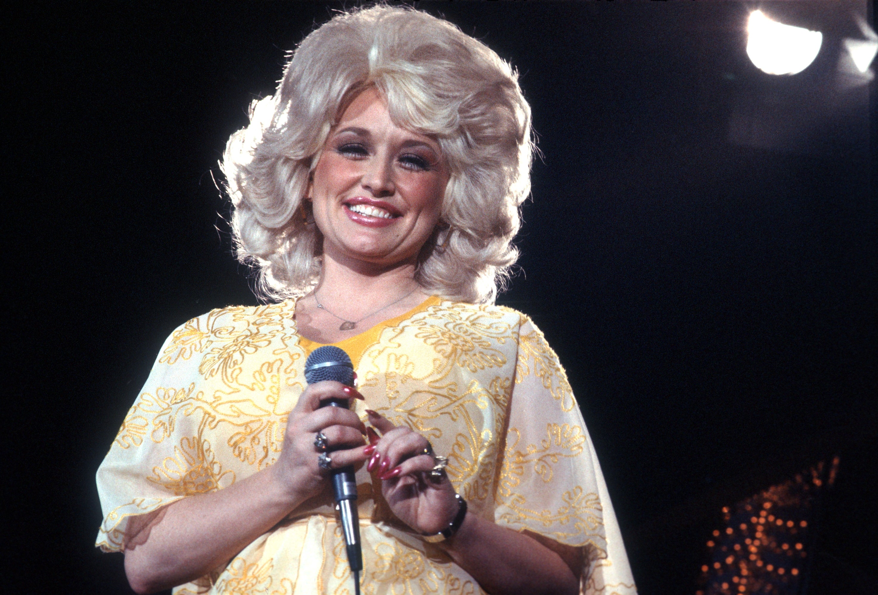 A person stands smiling while holding a microphone, wearing a floral-patterned outfit with voluminous hairstyle on stage